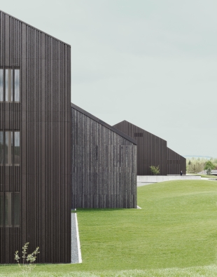 Das architektonische Konzept zitiert den Archetypus des Gehöfts aus einzelnen und in Gruppen stehenden Satteldachhäusern. Die neuen Trakte sind mit unterschiedlich gefalzten und individuell gekanteten Blechen mit 90°-Winkeln bekleidet. Die Bestandsbauten erhielten eine silbrig-graue Boden-Deckelschalung aus Holz. Die farblich aufeinander abgestimmte Bekleidung der VHF schafft ein stimmiges Gesamtensemble. Foto: Brigida González