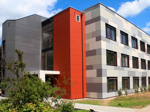 Ein kräftiges Rot setzt im Eingangsbereich einen warmen Akzent zur vorgehängten hinterlüfteten Fassade (VHF) des Gebäudekomplexes in Grau-Nuancen Foto: FRAHAMMER GmbH & Co. KG